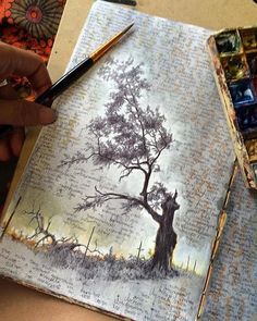 a person is holding a paintbrush and drawing on an open book with a tree in the background