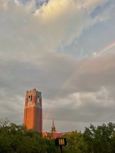 a tall tower with a rainbow in the sky above it and trees around it on a cloudy day