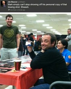 a man sitting at a table with a beer in front of him and people standing around