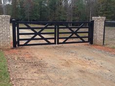 a black gate with brick pillars on the side of a dirt road in front of trees