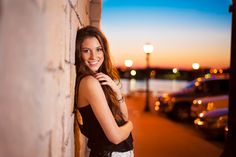 a beautiful young woman leaning against a brick wall at night with the city lights in the background