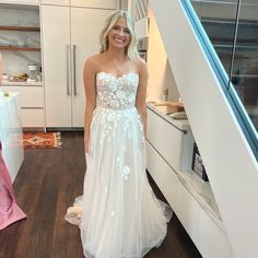 a woman in a white wedding dress standing next to a kitchen counter and stairs with two women looking at the camera