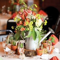 an arrangement of flowers in a watering can on a table with wine glasses and utensils