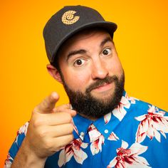 a man with a beard wearing a hawaiian shirt and hat pointing at the camera while making a funny face