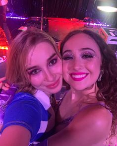 two young women taking a selfie in front of a stage with bright lights and music equipment