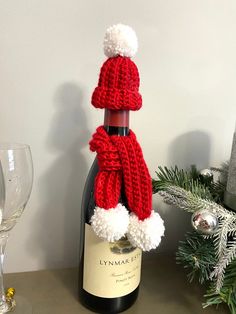 a bottle of wine with a knitted hat and scarf on it next to a christmas tree