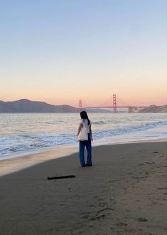 a woman standing on top of a sandy beach next to the ocean with a bridge in the background