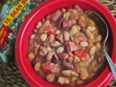 a red bowl filled with beans and ham on top of a woven table cloth next to a bag of beans