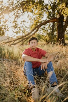 a young man sitting in the grass with his hands on his knees looking at the camera