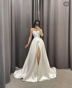 a woman taking a selfie in front of curtains wearing a white dress with thigh high slit