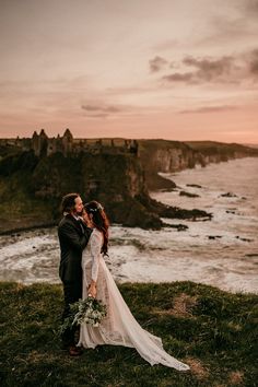 a bride and groom standing on top of a grass covered hill next to the ocean