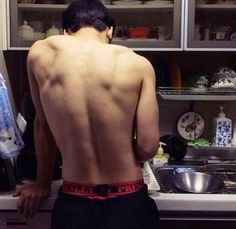 a man with no shirt standing in front of a sink and looking at dishes on the counter