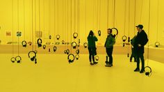 three people standing in front of a yellow wall with headphones hanging from the ceiling