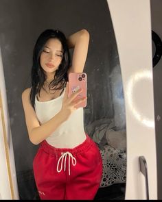 a woman taking a selfie in front of a mirror wearing red shorts and a white tank top