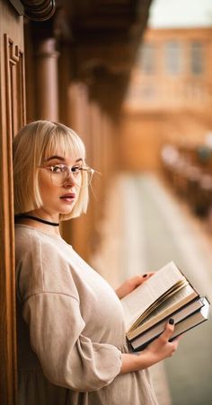 a woman with glasses is leaning against a wall and holding a book in her hands