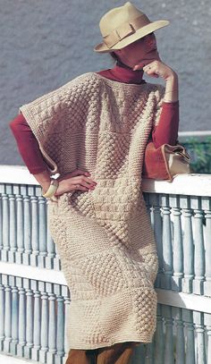 a woman wearing a knitted sweater and hat leaning against a fence with her hand on her chin