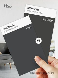 The image compares two dark paint swatches. On the left is "Graphite" by Benjamin Moore with the code 1603, a rich, deep charcoal gray with neutral undertones. On the right is "Iron Ore" by Sherwin Williams with the code SW 7069, a dark charcoal with warm undertones. A hand is holding both swatches, and the background is blurred, featuring a neutral-toned room. These colors are ideal for creating dramatic, modern spaces or accent features. Ship Lap Walls