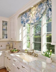 a kitchen with white cabinets and marble counter tops in front of a window that has blue floral curtains