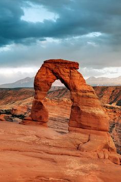 an arch shaped rock formation in the desert under a cloudy sky with mountains in the background