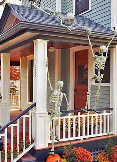 two skeleton statues on the porch of a house with pumpkins and flowers in front