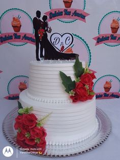 a white wedding cake with red roses on the side and a bride and groom topper