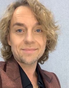 a man with blonde hair wearing a brown suit and black shirt smiling at the camera