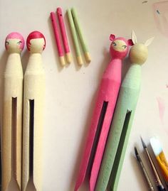 three wooden peg dolls are lined up next to markers and pencils on a white surface