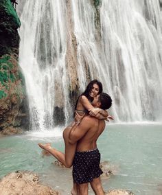 a man and woman hug in front of a waterfall while they stand on rocks near the water