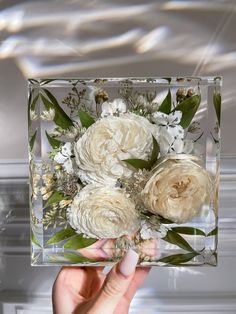 a hand holding a clear square vase with white flowers and greenery on the inside