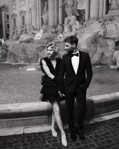 a man and woman standing next to each other in front of a fountain wearing tuxedos