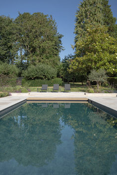 an empty swimming pool surrounded by trees and bushes, with two lawn chairs in the background