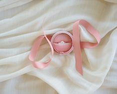 an open ring box with a pink ribbon around it on top of a white sheet