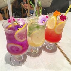 three glasses filled with different colored drinks on top of a white table covered in flowers