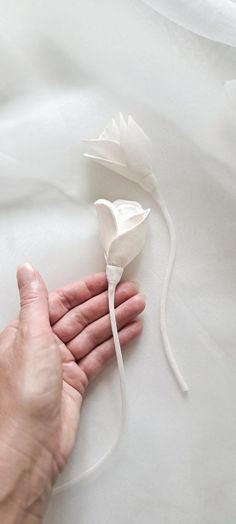 a person's hand holding a white flower on top of a bed with white sheets