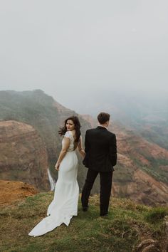 a bride and groom standing on top of a mountain looking out at the valley below