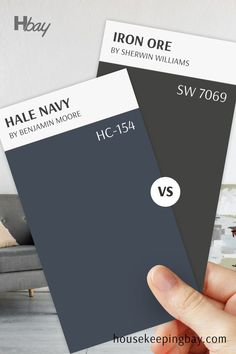 The image compares two dark paint swatches. On the left is "Hale Navy" by Benjamin Moore with the code HC-154, a deep navy blue with cool undertones. On the right is "Iron Ore" by Sherwin Williams with the code SW 7069, a dark charcoal gray with warm undertones. A hand is holding both swatches, and a "VS" symbol in the middle indicates a comparison. The background is blurred, featuring neutral interior elements. These colors are bold and versatile, ideal for accent walls or exterior features. Sherwin Williams Coordinating Colors, Hale Navy