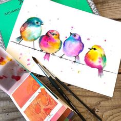 three colorful birds sitting on top of a wooden table next to watercolors and paintbrushes