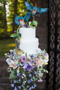 a three tiered white wedding cake with blue butterflies on top and flowers around it