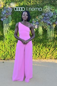 a woman in a pink one shoulder jumpsuit standing next to a wall with succulents