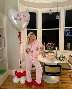 a woman in pink pajamas standing next to a birthday cake and balloon with the words happy birthday written on it