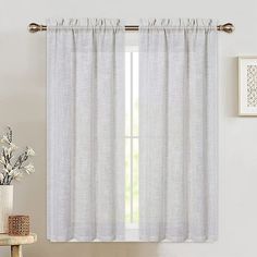 a white curtain hanging on the side of a window next to a chair and vase with flowers