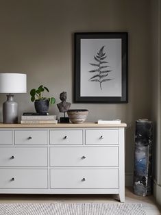 a white dresser with two vases on top of it next to a framed plant