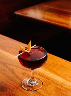 a glass filled with red liquid sitting on top of a wooden table next to a slice of orange