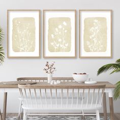 three framed art prints on the wall above a dining room table with white chairs and a plant