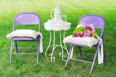 two purple chairs and a white table with flowers on it are sitting in the grass