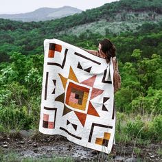 a woman holding up a quilt in the woods with mountains in the backgroud