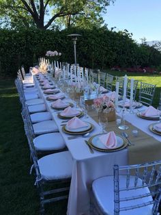 a long table is set with white and gold place settings for an outdoor wedding reception