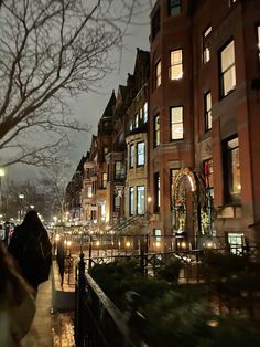 people walking down the street at night in front of some very tall buildings with lights on them