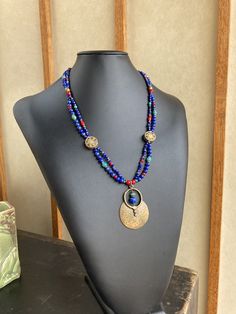 Featuring stones that are associated with wisdom, insight, spirituality, and communication, this is made with gorgeous deep royal blue to navy blue grade A Lapis in various shapes and sizes, which contrast strikingly with green semi precious turquoise, rich red Czech glass and white-heart beads, and brass spacers. The two strands gather at Tibetan brass 15mm coin beads inlaid with red coral and turquoise howlite as well as other brass beads. A 53x37mm plated antique gold large tribal style medal Artisan Blue Necklace With Gemstone Beads, Bohemian Blue Lapis Lazuli Beaded Necklace, Bohemian Blue Gemstone Bead Necklaces, Blue Lapis Lazuli Jewelry For Festivals, Handmade Bohemian Sapphire Necklace, Bohemian Lapis Lazuli Beaded Necklaces As Gift, Bohemian Lapis Lazuli Beaded Necklaces For Gift, Blue Amulet Style Necklace With Natural Stones, Blue Amulet Necklaces With Natural Stones