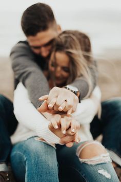 a man and woman holding hands while sitting on the ground in front of an ocean